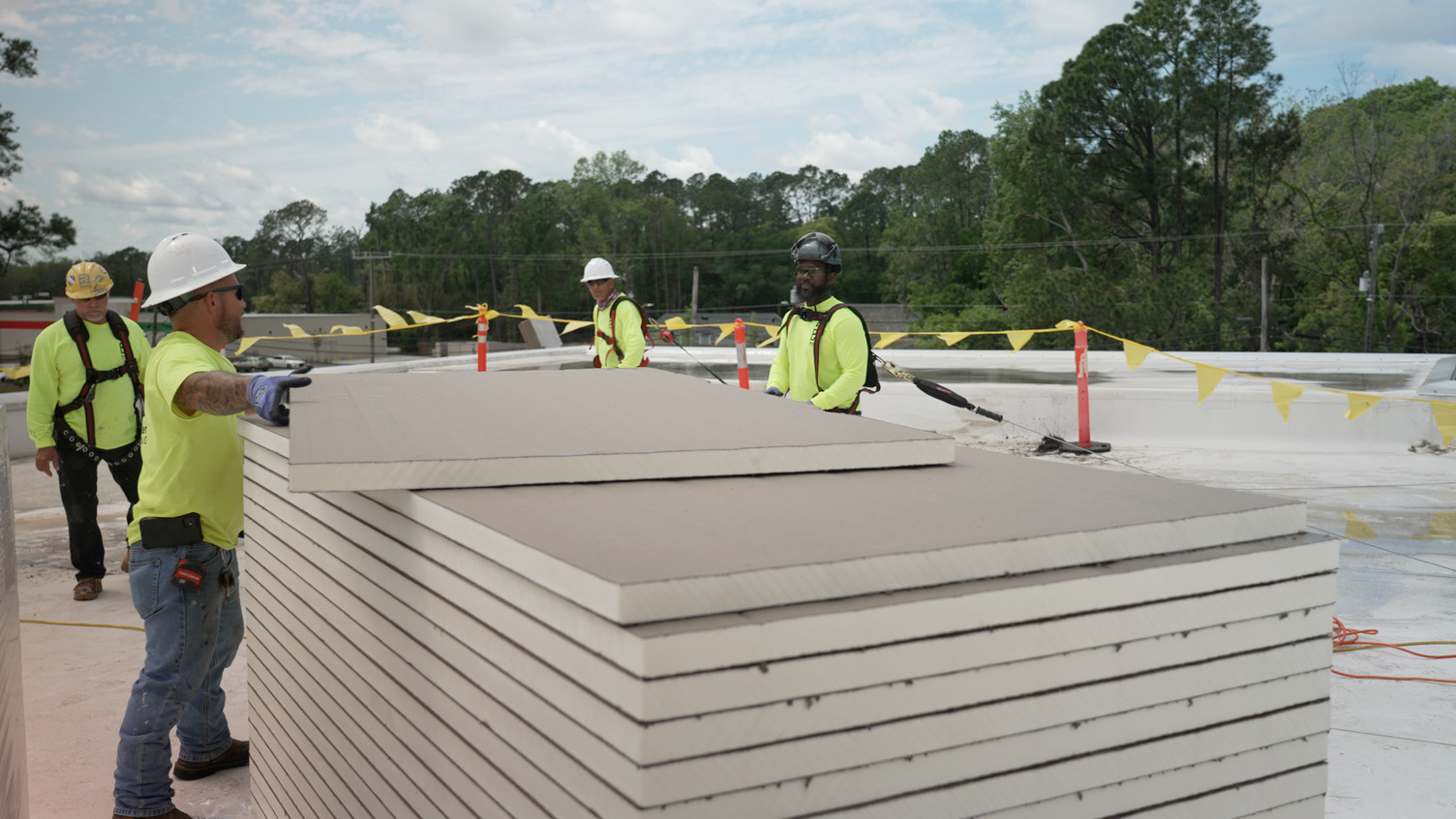 Four construction workers in safety gear handle a large stack of insulation boards on a commercial flat roof